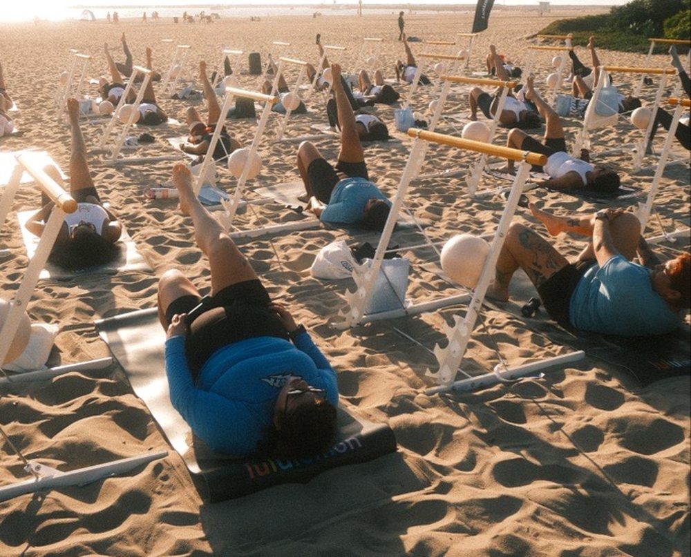 A barre class on the beach in golden light, with people doing leg lifts at rows of white barres
